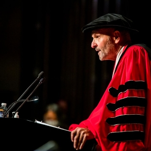 UArts President David Yager, seen squarely from his left, is dressed in a crimson doctoral robe emblazoned with black stripes on the arm, speaks into two microphones at a black podium.