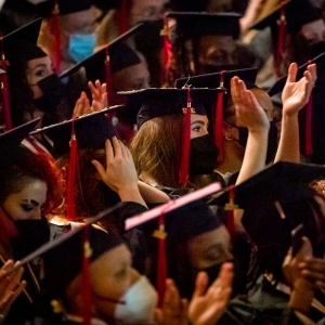 a dense, seated crowd of people in black graduation garb are seen from the side, clapping and looking ahead. Their tassels are red and on the right side of their graduation caps. All are wearing face masks, mostly black ones. The person in the center of the image and most in-focus is holding their hands higher than anyone else around them. 