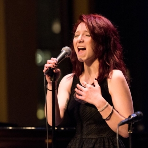 a person with long red hair in a black dress sings into a mic with their eyes closed. the background is out of focus, but reflected stage lights and a possible closed piano lid are implied. 