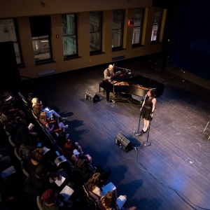 a vocal recital is seen from above. to the left are several rows of chairs with an audience facing towards the right of the image, where a single singer in a black dress sings into a microphone. a person in a brown suit vest plays on a grand piano towards the far wall, the windows of which reveal that the recital room is many floors up in a dense city. 