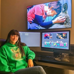 Emily Angelucci sits at a desk with her left elbow on the desk, turned towards the viewer and smiling. she is wearing a green hoodie. on the desk is a double-monitor video editing setup, showing a detailed project being cut. 