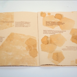 an open book without a jacket or cover. the pages are a beige tone and jagged at the edges. the pages are stamped or inked with pentagonal sepia ink splotches. brown text prose interplays across the pages