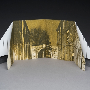 a folded paper structure with a printed image depicts a gate between two buildings rendered in sepia. founded paper elements prop up this gate from the sides, adding depth with a printed brick texture, extending the imagined alley further forward.