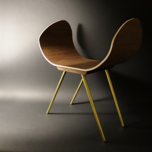 The image appears to be a 3d rendering but it could just as easily be a minimal photograph. A curved seat rendered in a warm wood grain is set atop four brass-like legs diagonally angled below its base. The overall shape of the furniture is inviting.