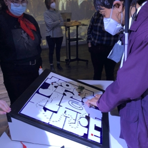 A person in a purple jacket is leaning over a tablet interface, displaying a map of a museum interior with tangible waypoints. 