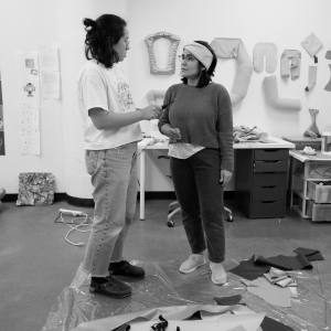 Two people stand and converse in the center of a large studio space with white walls covered in art and sketches. The person on the left is wearing jeans, a white shirt, and long hair in a loose bun seen in profile, while the person on the right is wearing a sweater, dark jeans, and a scarf around their forehead. The floor is covered in scraps of textiles on a large plastic covering. There is a desk against the studio wall, above which are many bits of artwork pinned to the walls. Image is black and white.