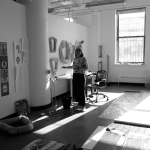 A person stands next to a desk in a large, open studio as sunlight streams in. the left wall is white drywall covered in artworks, interrupted by a concrete pillar. The right, back wall has large windows that allow large beams of light to fall across the floor, which is covered in textiles and plastic sheeting. The person is wearing a floral shirt and is smiling. Image is black and white.