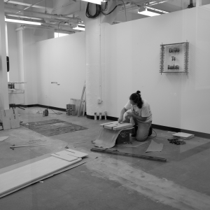 A person wearing a mask in a large, empty studio space is down on one knee working on an artwork - a square canvas resting on a milk crate. There are various sheets of material and wooden elements scattering across the floor. The wall behind the person is largely blank, with one artwork reading “Dios te bendiga.” The image is overlaid with reflections, implying that the photo is shot through an interior window into a hall. Image is black and white.