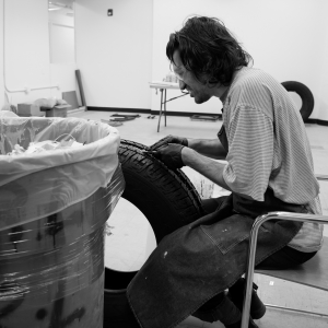 A person in a large striped shirt and apron is seen in profile, seated in a largely empty studio space with blank white walls. The person has safety goggles and black latex gloves on as they perform work on a large black vehicle tire held between their knees. A gray trash can occupies the left foreground of the image, obscuring half of the tire. Image is black and white.