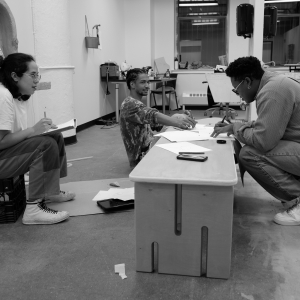 Three people sit low to the floor in a studio space. One person on the left is seen in profile writing on a notepad, sitting on a milkcrate. Two people in the center of the image are writing on sheets of paper on a slim, low wooden table. The person in the center of the group is sitting down and looking at the person on the right, who is crouching on the balls of their feet. 