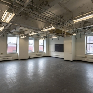 A large, well-illuminated room with white walls, a slate gray floor, and an exposed ceiling full of rows of pipes and fluorescent lighting fixtures. Several windows are on the far walls, letting in light from the cityscape outside. On the right wall is a black television screen and a window with protective bars. 