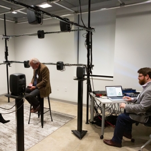 Two people sit on high stools within a loose scaffold supporting an array of small, high-grade speakers. Both people are looking at their phones or notebooks. A person on the right of the image sits at a desk with a laptop displaying a schematic, ostensibly controlling the speaker array via a dense collection of cables spilling out. 