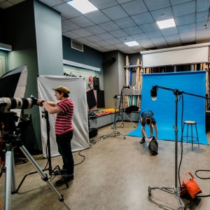 A photography studio full of equipment and people at work. A blue seamless drop background is in the center towards the back of the paneled room. A few lights on boom stands can be seen throughout the room, with several people handling equipment all around. A person by the seamless is bent over reaching into a bag, a person on the right is squatting and handling cables. A person on the left is standing in profile, adjusting a light. 