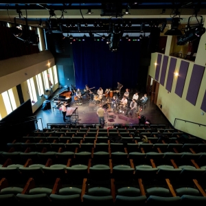 Interior of a recital hall or musical performance space. The viewer’s perspective is from far up in the seating area, looking down towards the stage area across eight rows of folding green theater seating. In the stage area, an instrumental ensemble is practicing.  