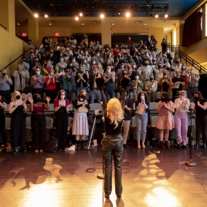 a view of caplan recital hall from the stage. a person with long blond hair and leather pants faces the audience with both hands in the air. the rakes auditorium seating is packed with people who are all standing and interacting with their hands, either clapping or raising them. people are wearing face masks. 