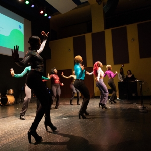 a group of dancers in brightly colored tops and tight pants are dancing on large high heels on the floor of caplan recital hall. the view is from stage right looking towards the dancers in low lighting, with acoustic paneling seen against the yellowish rear wall and a drum kit atthe back of the stage on the left. a projection above the event on the image's left reads "excel" in green blocky type. 