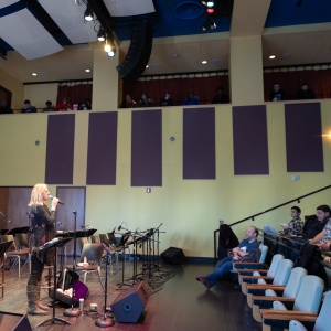 a person with blond hair and tall boots is seen in profile among band seating on the stage of caplan recital hall, speaking into a microphone addressing a crowd seated in the theater seating toward the right. 