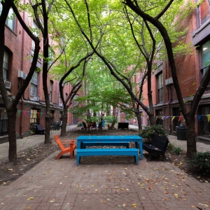 a view of furness courtyard from within, looking out towards 15th street. in the foreground is a large blue plastic picnic table with matching benches and an orange plastic lawn chair. twisting trees ring the seating areas, themselves ensconced by the dark red brick walls of the building. 