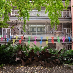 a view from within furness courtyard looking into hamilton hall's atrium. a rusted metal sculpture composed of waves of triangles is in the foreground, flanked by ginko trees whose bright green leaves hang from above. multi-colored pennants span between the two trees.