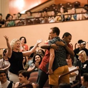 interior of levitt auditorium, with a dynamic image of the audience during a new student orientation event. some people are seated in the seating area as well as in the upper floor balcony, while a number are standing, clapping, and embracing. 
