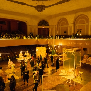 interior shot of levitt auditorium, which is bathed in golden light. the chamber's ceiling is ornate, with a large chandelier suspended above the crowd milling about on the auditorium floor where a number of sculptures and installations are displayed.