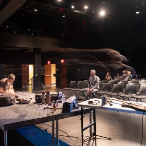 a view from the stage of caplan studio theater's black box during setup for a performance. a number or scattered instruments and tools are lying on folding tables on or near the stage, including electric guitars and shakers. a few people are in the seating. the ghostly image trail of a person is soon from a long exposure at the right of the image