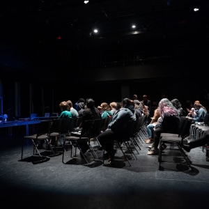 an image of the seating during the Out Of The Box performance in the caplan studio theater. the seating is seen in profile from ithe left of the audience, and the chairs are all on the flat floor - not raked. the audience is illuminated with white light while the empty stage is washed in blue light. 