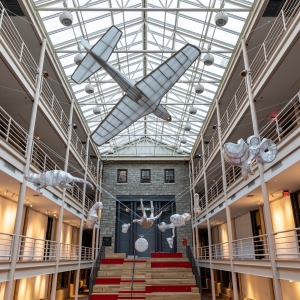 interior of solmssen court, looking upwards toward the glass roof, behind which is a gray sky. the entire chamber is devoid of people. warm lights illuminate the walls on the first and second story walkways. several sculptures are suspending in mid-air, including a very large airplane model, a human figure, abstract shapes, and a rat-type critter.