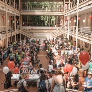 interior of solmssen court during an event. the view is from the living steps, looking down into the ground floor, which is ringed by walkway balconies. warm, summer light floods in from above. rd and white balloons float from all of the railings. the event below consists of people in color-coded tees sitting at folding tables with various materials available on display.