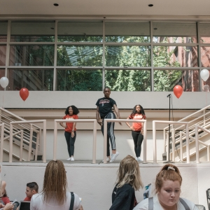 dancers performing on the stair landing in solmssen court between the ground second floor levels. a person in a black shirt and black joggers is towards the front, with two people with long black hair in red shirts and black pants flanking towards the back. red and white balloons float above the railings. 