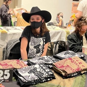  a person ins a black t shirt, black face mask, and black cowboy hat with long hair is seated at a table displaying several stacks of the same shirt they are wearing. people all around are seated at or walking past similar tables displaying printed artwork. 