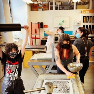 a group of students in the lithography studio. a person in the front left with a massive wallet chain and messy hair is holding a roller straight overhead. in the center, a person with long hair looks down and wets a stone with a sponge, a metal bowl in the other hand. a person in the far back is adjusting a press. all four people are wearing face masks. 