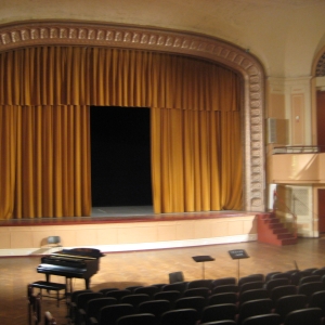 interior of levitt auditorium. the auditorium is empty, seen from the top of the central seating area. golden-yellow curtains are hanging across the large stage, with a large rectangular rift left open.  