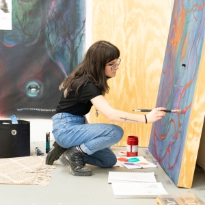 a fine arts student in blue jeans is seen in profile kneeling and painting orange squiggle patterns on a large scale canvas on the floor. 