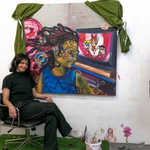a fine arts student sits at their canvas depicting a person with a molten face in a blue top and a power puff girls episode in the background. the painting is surrounded by green drapes. 
