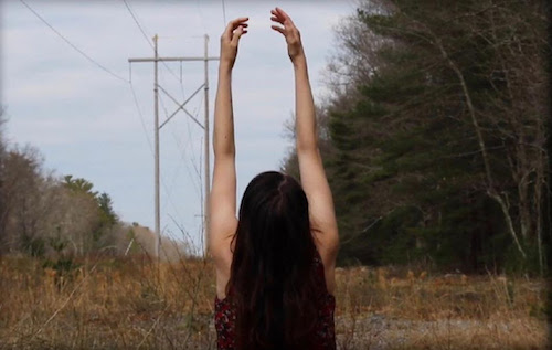 A dance student raises their arms to the sky in a field with trees and a telephone pole.